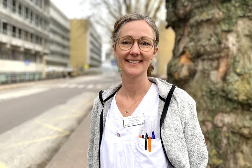 Margret Leosdottir med sjukvårdskläder, namnskylt och pennor i fickan står utomhus vid ett träd och en byggnad och tittar in i kameran.