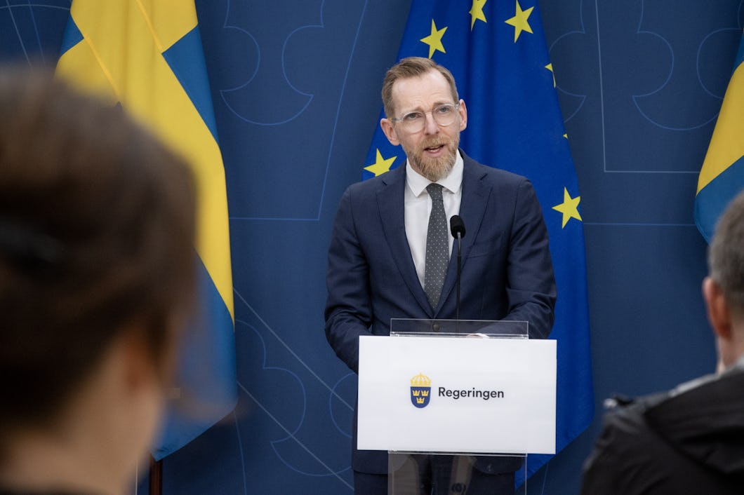 Socialminister Jakob Forssmed i kostym talar vid en talarstol märkt "Regeringen", med svenska och EU-flaggor i bakgrunden.