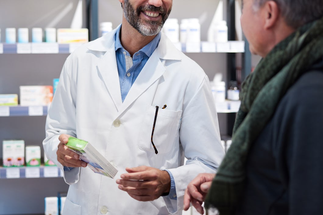 En farmaceut i vit rock håller i en medicinförpackning och samtal med en kund inne på ett apotek. I bakgrunden syns hyllor med olika produkter.