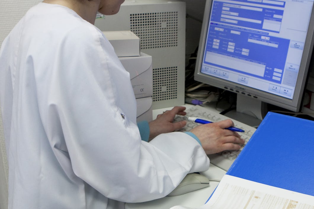 En person i laborationsrock matar in uppgifter i en dator vid ett skrivbord med en bildskärm, ett tangentbord och dokument.