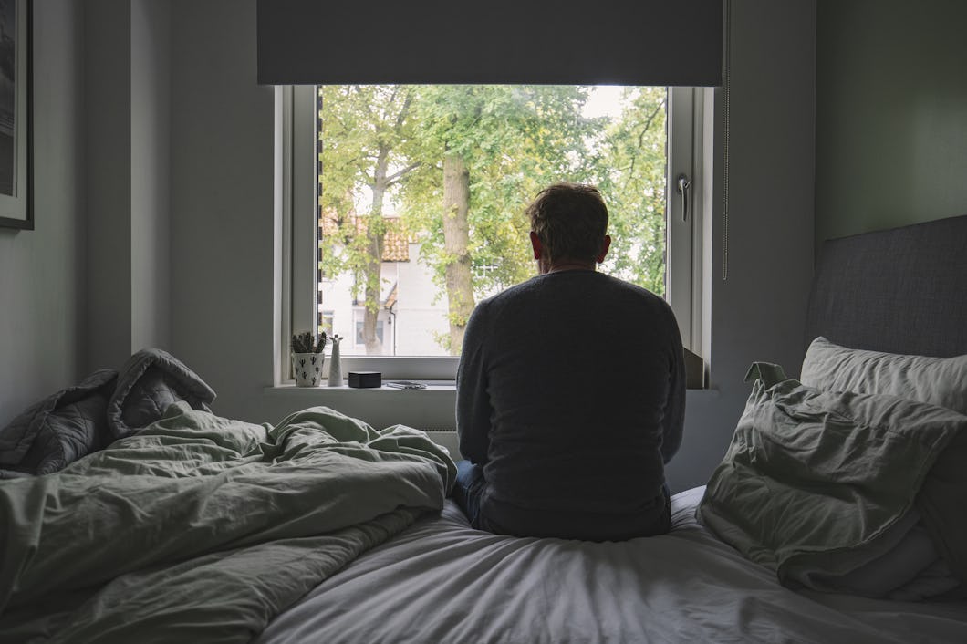 En person sitter på en säng vänd mot ett fönster med träd utanför, i ett sovrum där sängen inte är bäddad.