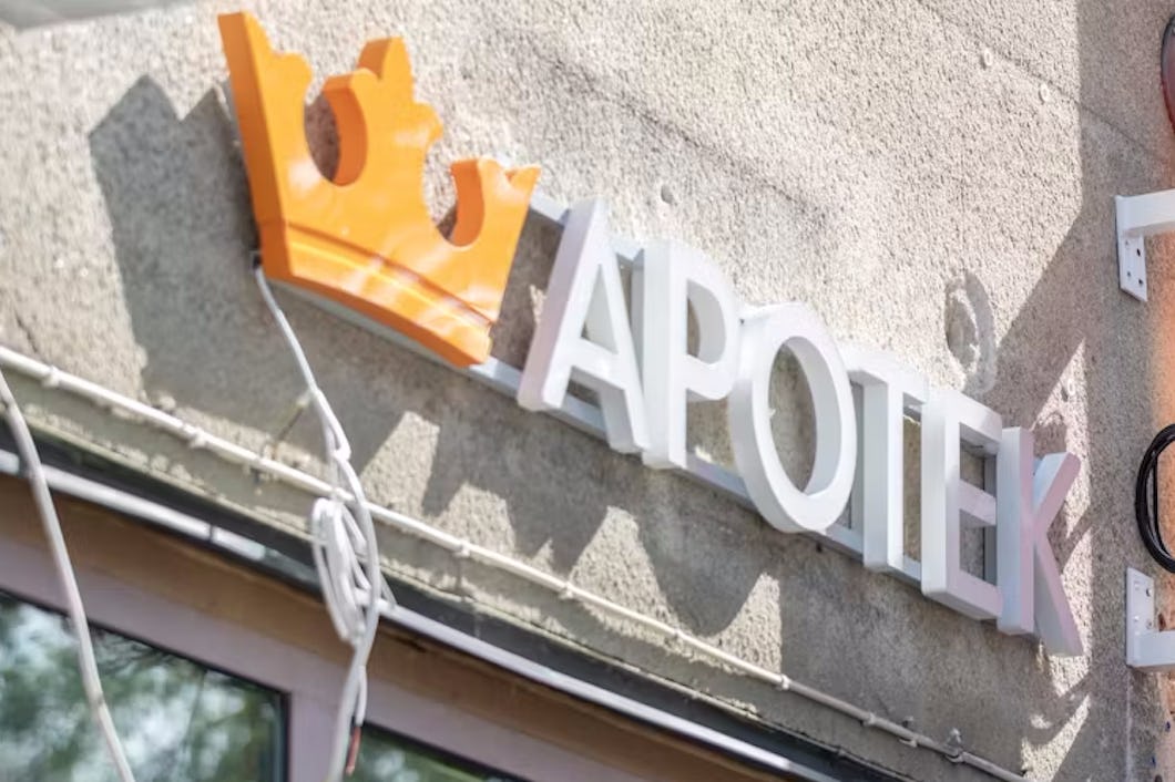 På utsidan av apoteket finns en skylt med texten "APOTEK" i vita bokstäver intill en orange krona. Skylten är monterad på en vägg av betong.