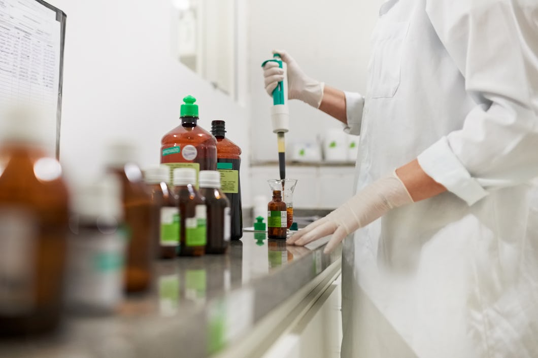 En person med labbrock och handskar använder en pipett för att överföra vätska till en brun glasflaska på ett laboratoriebord med flera flaskor.