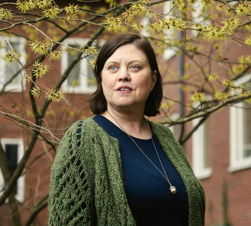 Katharina Erixon med kort brunt hår står utomhus utomhus och tegelbyggnad och ett träd med knoppar. Hon har på sig en mörkblå topp och en grönstickad kofta.