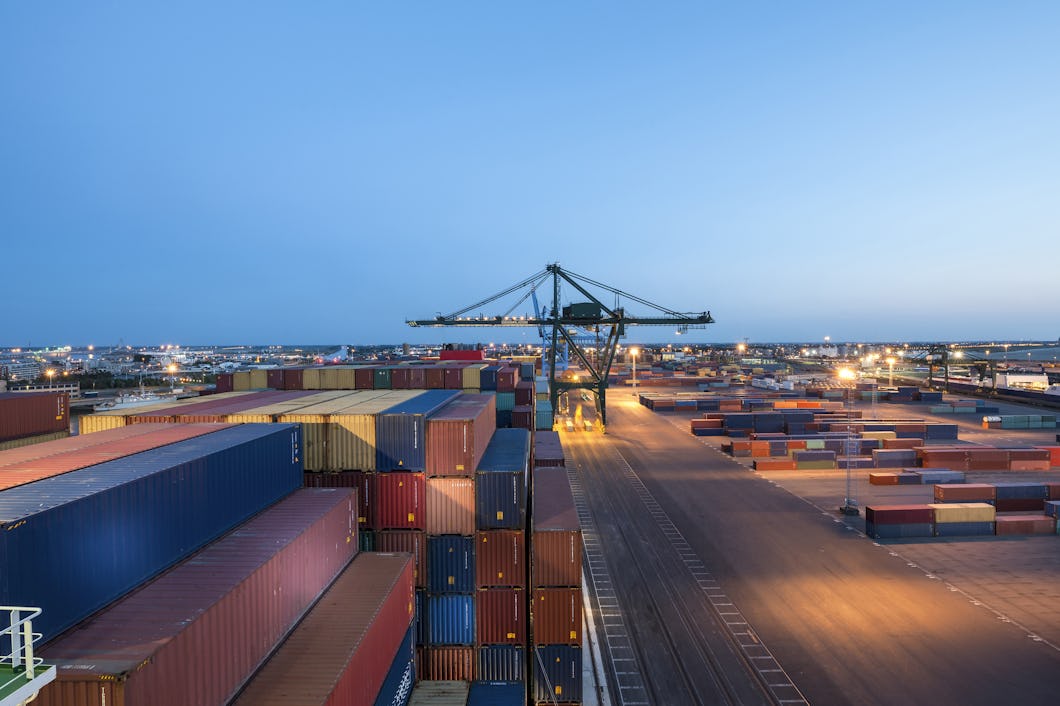 Containrar står staplade vid en hamnterminal, där kranar arbetar och området är upplyst under himlen.