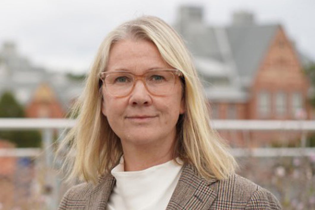 Karin Berglund med blont hår och glasögon står utomhus. Hon har på sig en rutig kavaj och en vit topp. I bakgrunden syns byggnader som inte är i fokus.