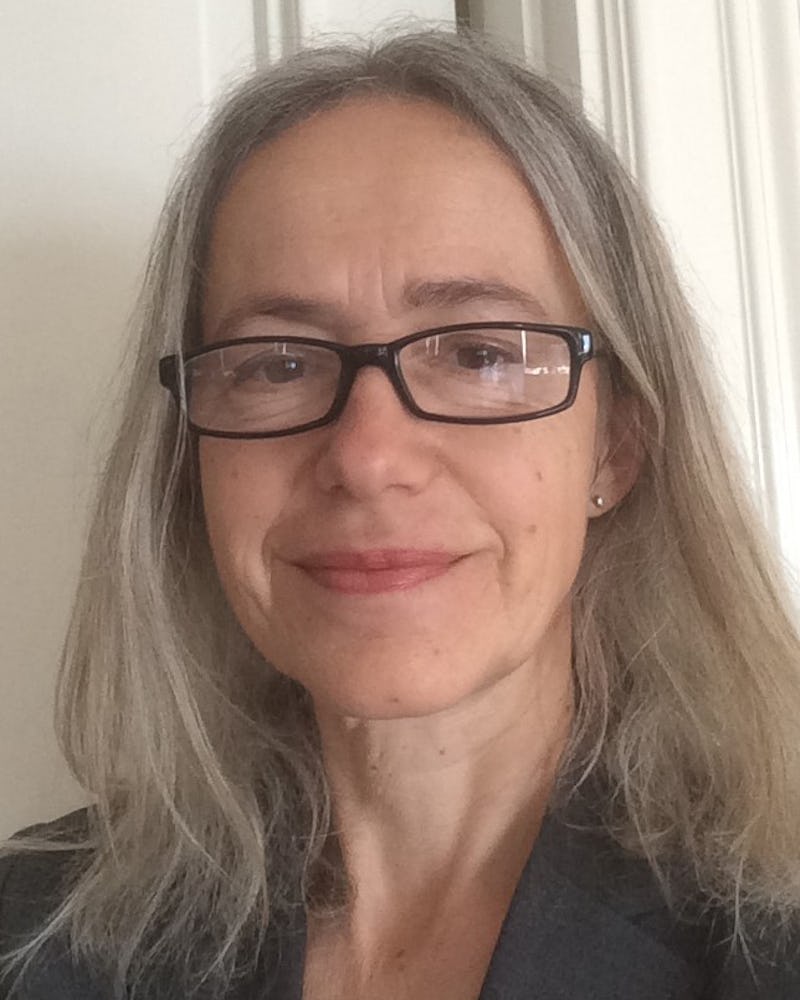 Anna Wettermark med långt grått hår, glasögon och en grå kavaj står inomhus framför en ljus vägg.