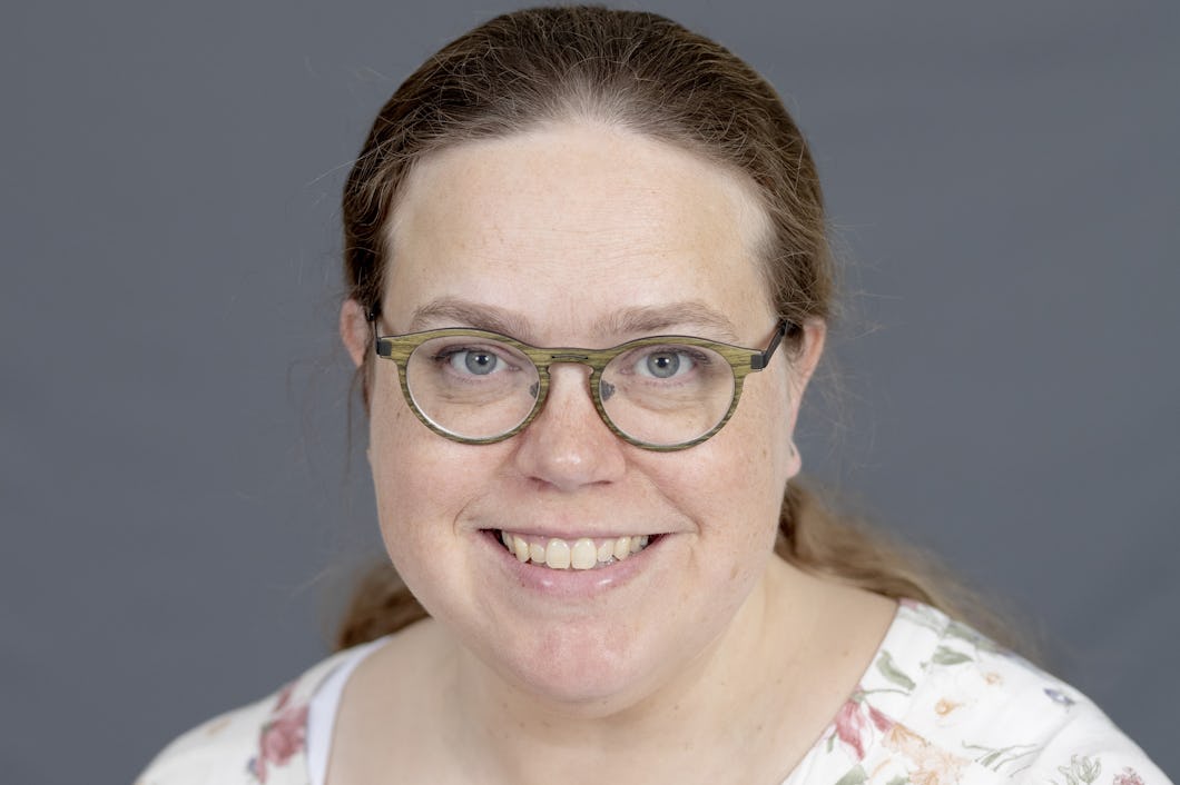 Christina Ljungberg Persson med långt brunt hår har runda glasögon och en vit topp med blommönster. Hon ler mot kameran framför en grå bakgrund.