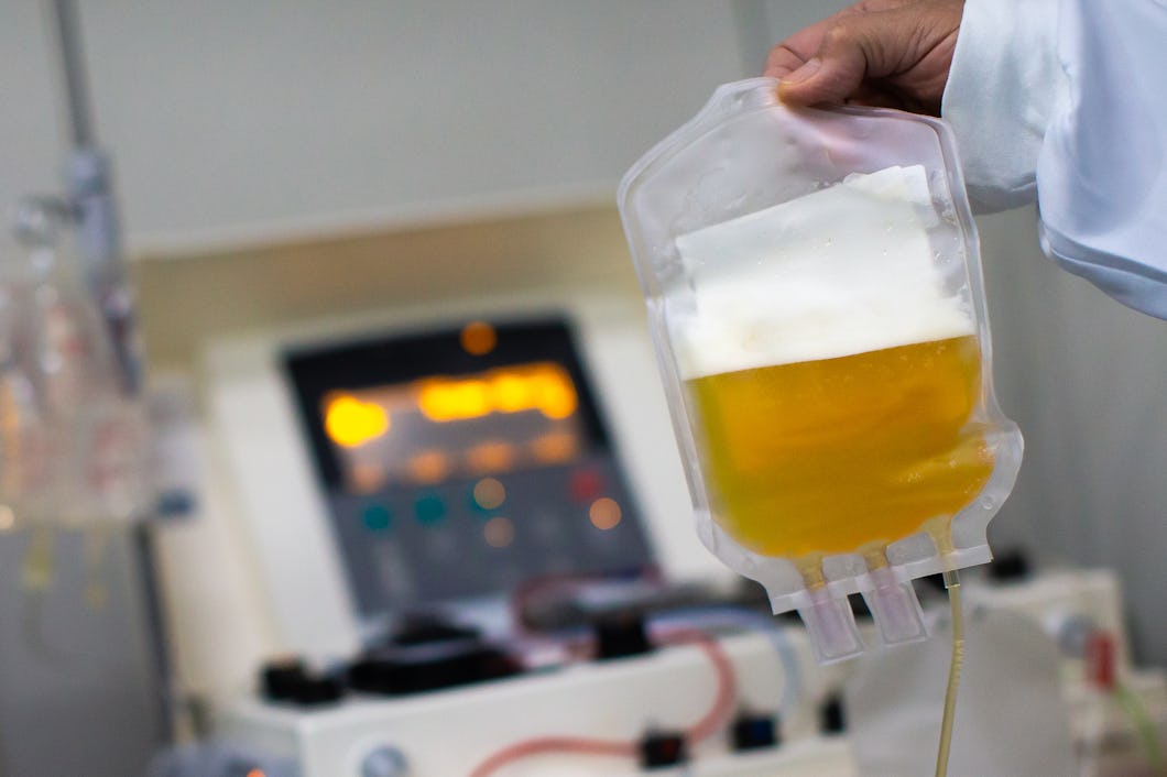 En person håller en medicinväska med plasma framför en medicinsk maskin i ett kliniskt rum.