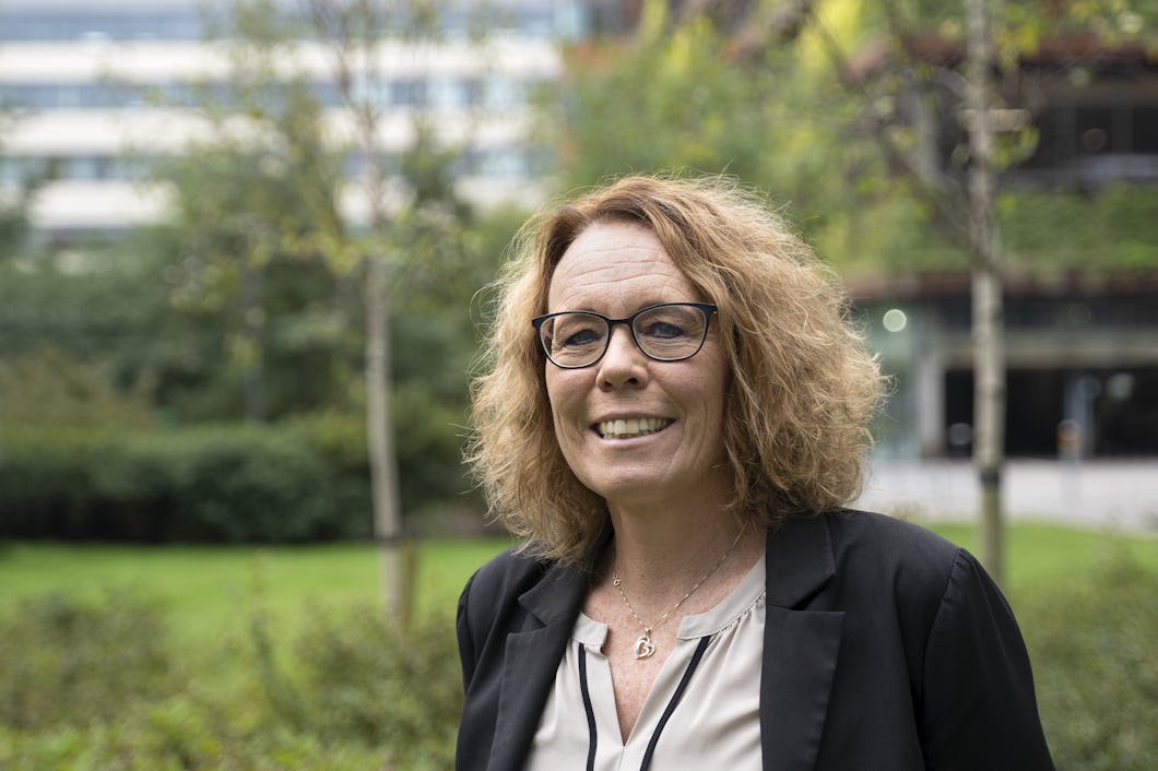 Maria Landgren med glasögon och lockigt hår står utomhus och ler. Hon befinner sig framför en gräsyta med träd och en byggnad i bakgrunden.