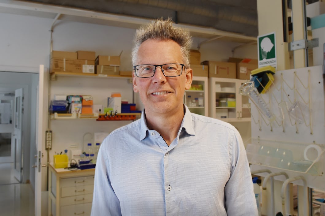 Tore Bengtsson med glasögon och ljusblå skjorta står i ett laboratorium med hyllor, utrustning och material i bakgrunden.