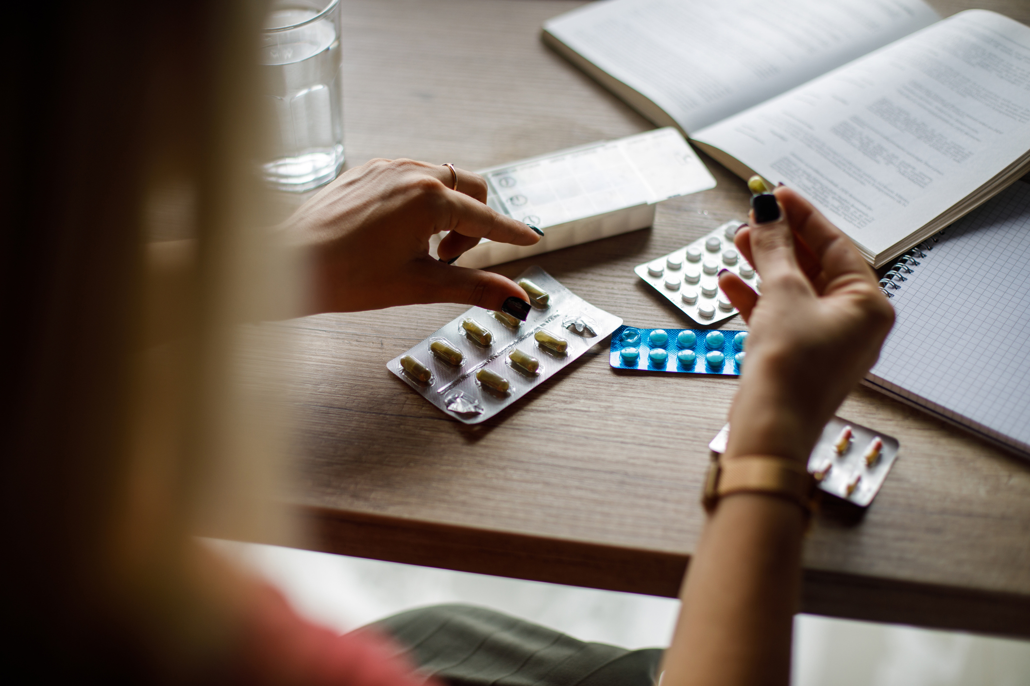 En person sitter vid ett skrivbord med tabletter i blisterförpackningar, ett glas vatten och en öppen anteckningsbok, håller en kapsel.