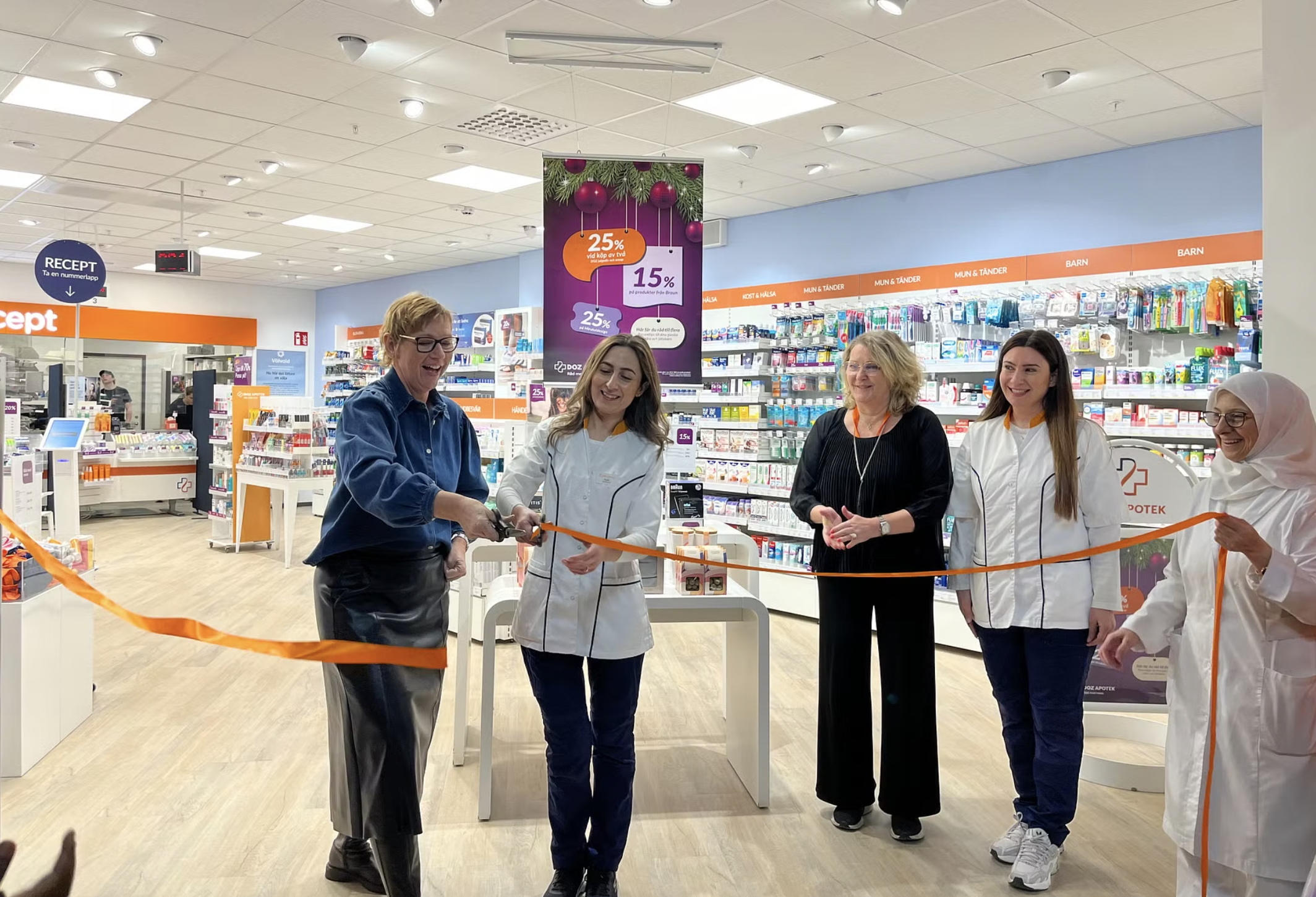 Fem kvinnor står i ett apotek, en klipper ett orange band vid en ceremoni. Produkthyllor och skyltar syns i bakgrunden.