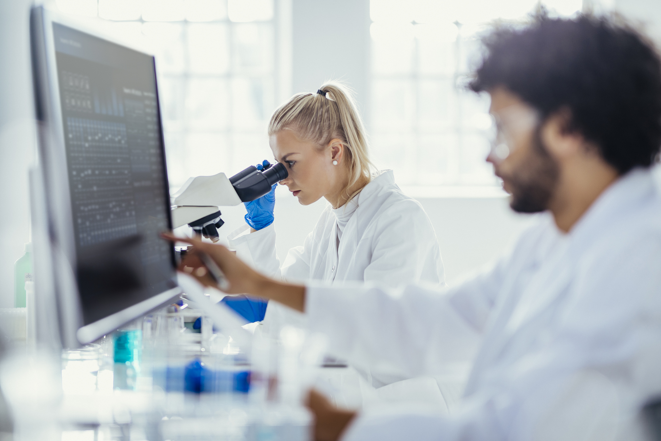 Två forskare i laboratorierockar arbetar vid skrivbord; en använder ett mikroskop och en granskar data på en datorskärm.
