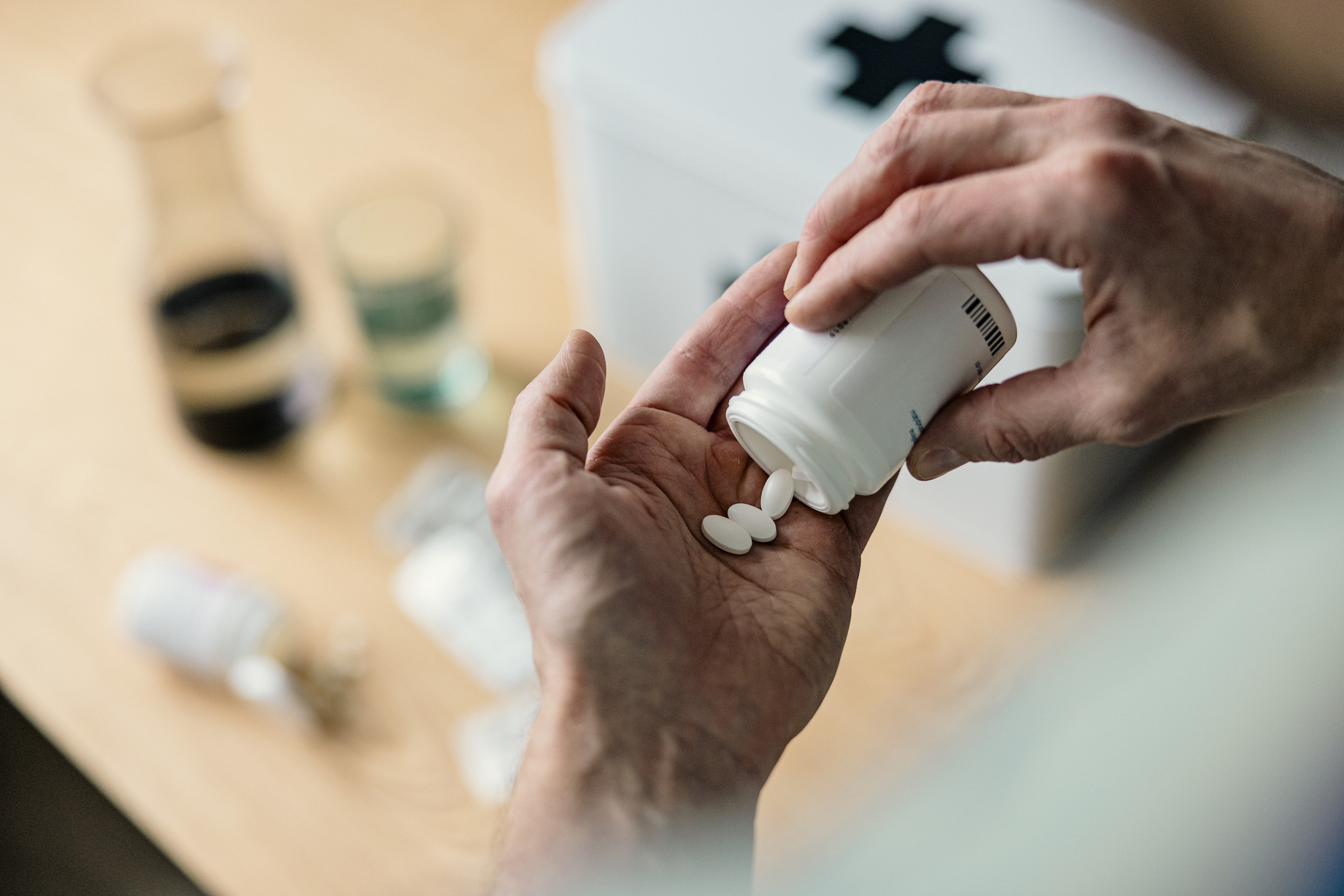 En person häller vita tabletter från en medicinflaska i handen, med medicinska förnödenheter och en vit första hjälpen-låda i bakgrunden.