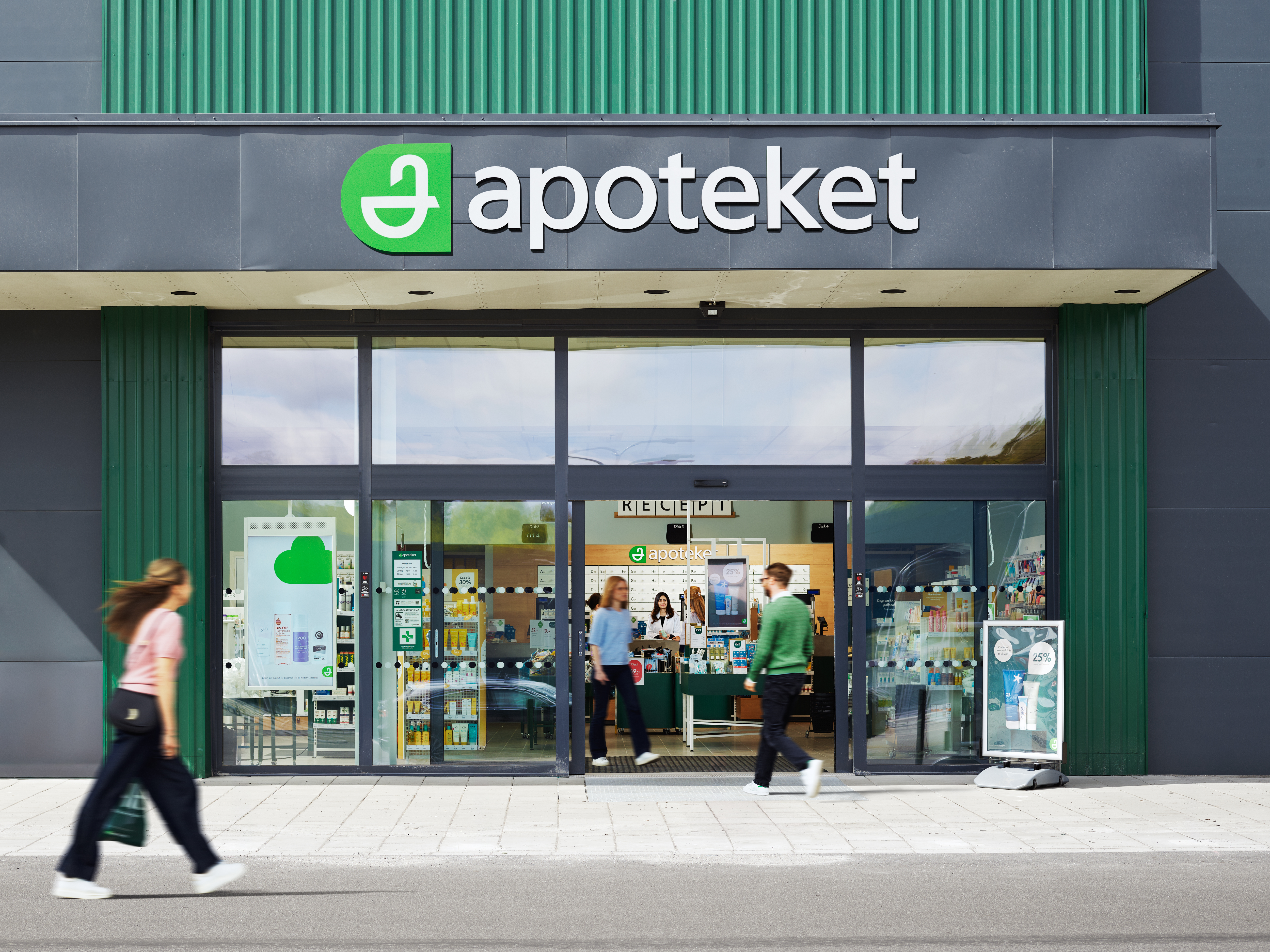 Huvudentré till ett Apoteket med glasdörrar, skylt ovanförför och personer som går förbi på trottoaren.