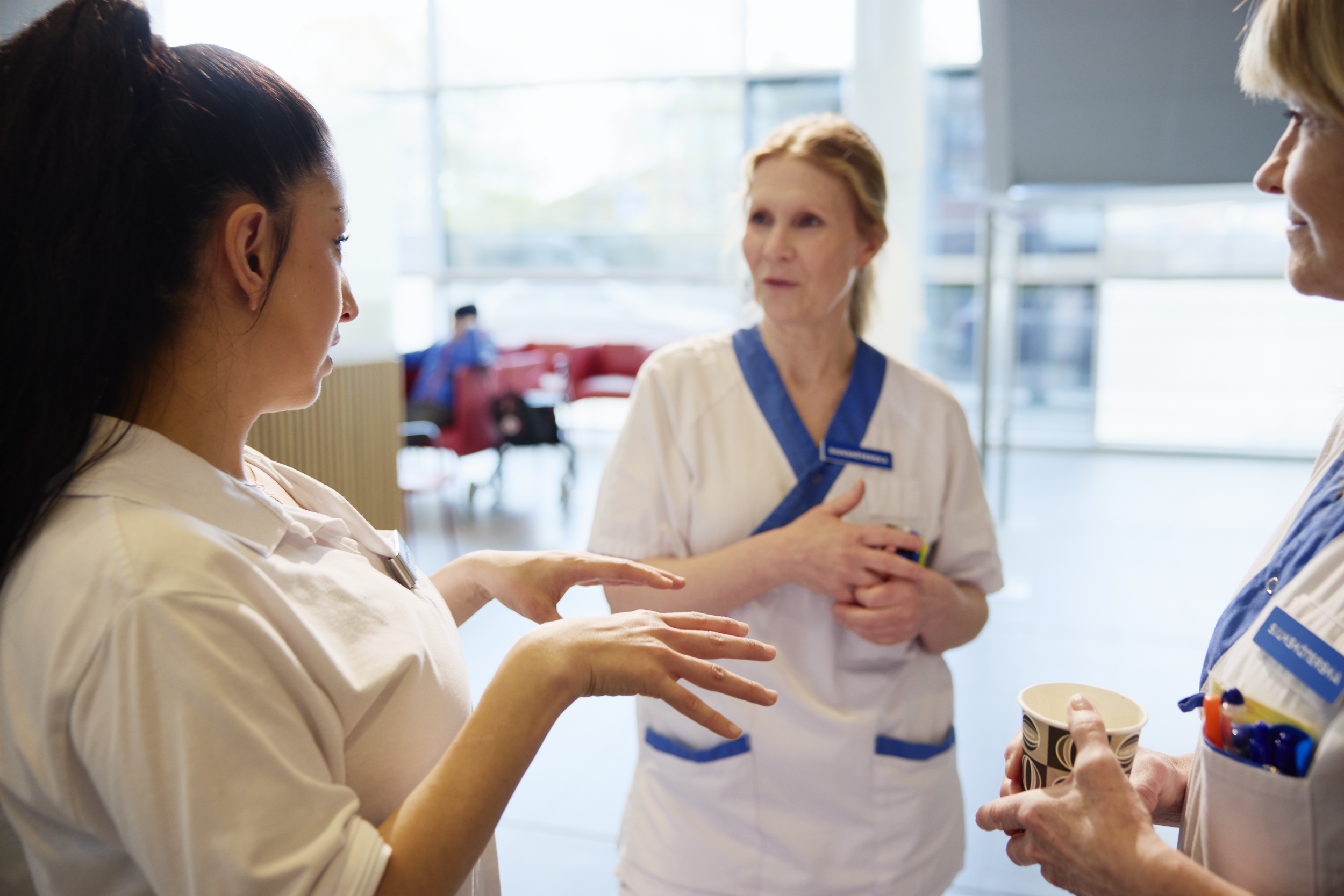 Tre vårdarbetare i arbetskläder står inomhus och pratar. En av dem håller en pappersmugg med kaffe.