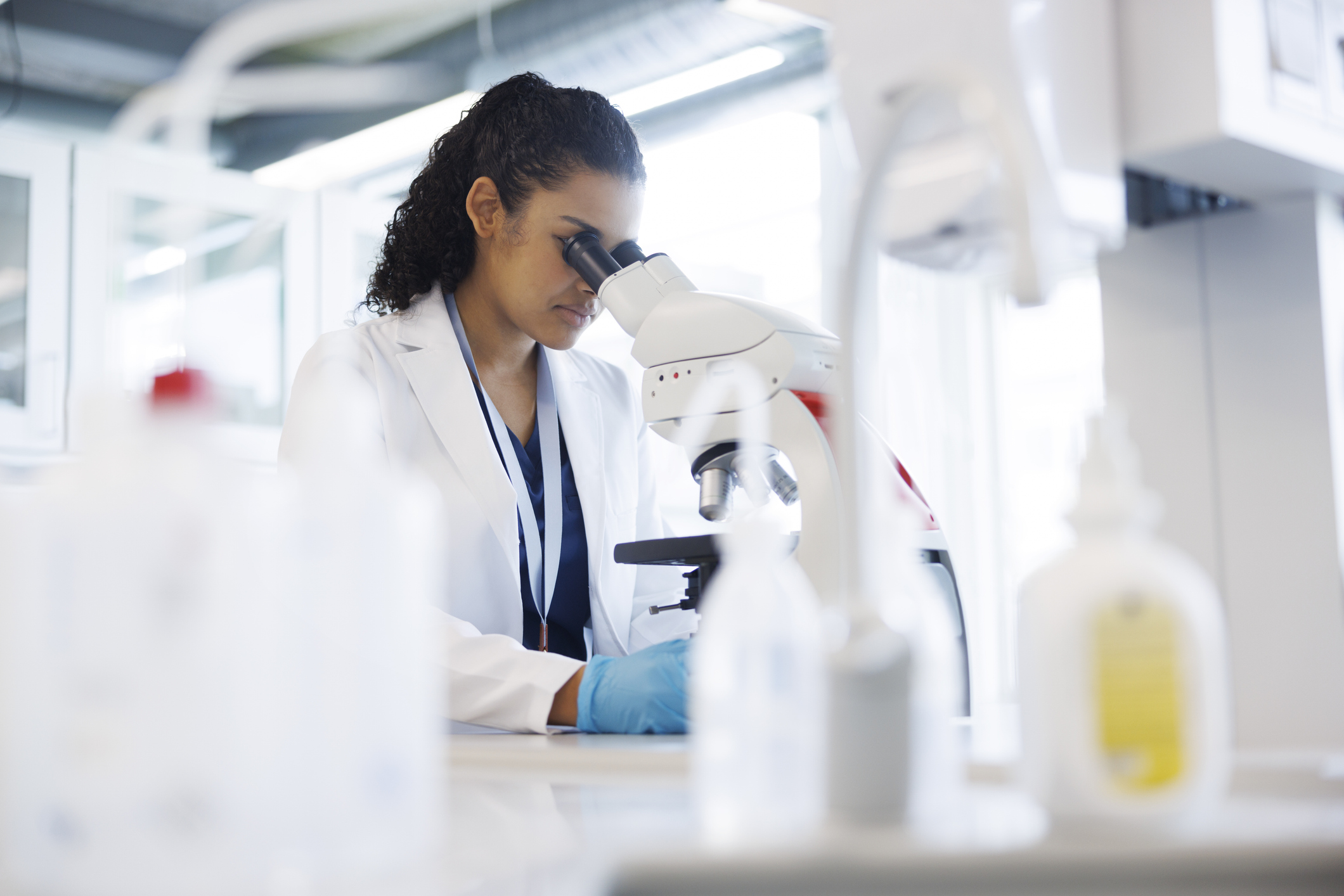 En forskare med laboratorierock och handskar tittar i ett mikroskop i ett laboratorium, med utrustning synlig i förgrunden.