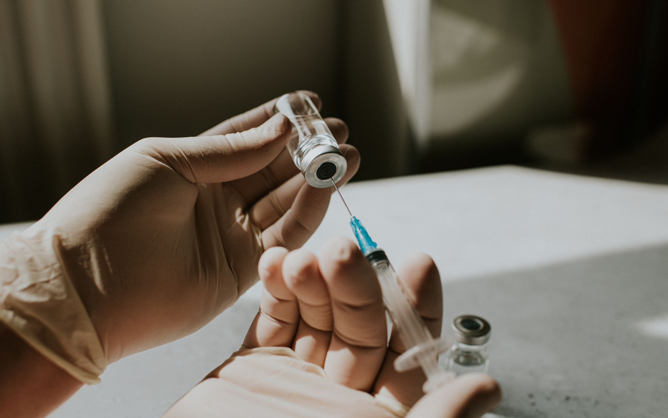 Handskar drar upp vätska ur en flaska med en spruta på en ljus yta i dagsljus.