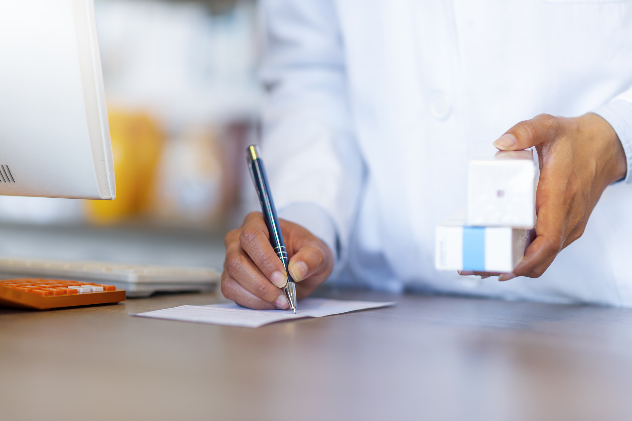 En person i vit rock skriver på papper vid ett skrivbord och håller två små lådor, med en dator och en orange kalkylator bredvid.