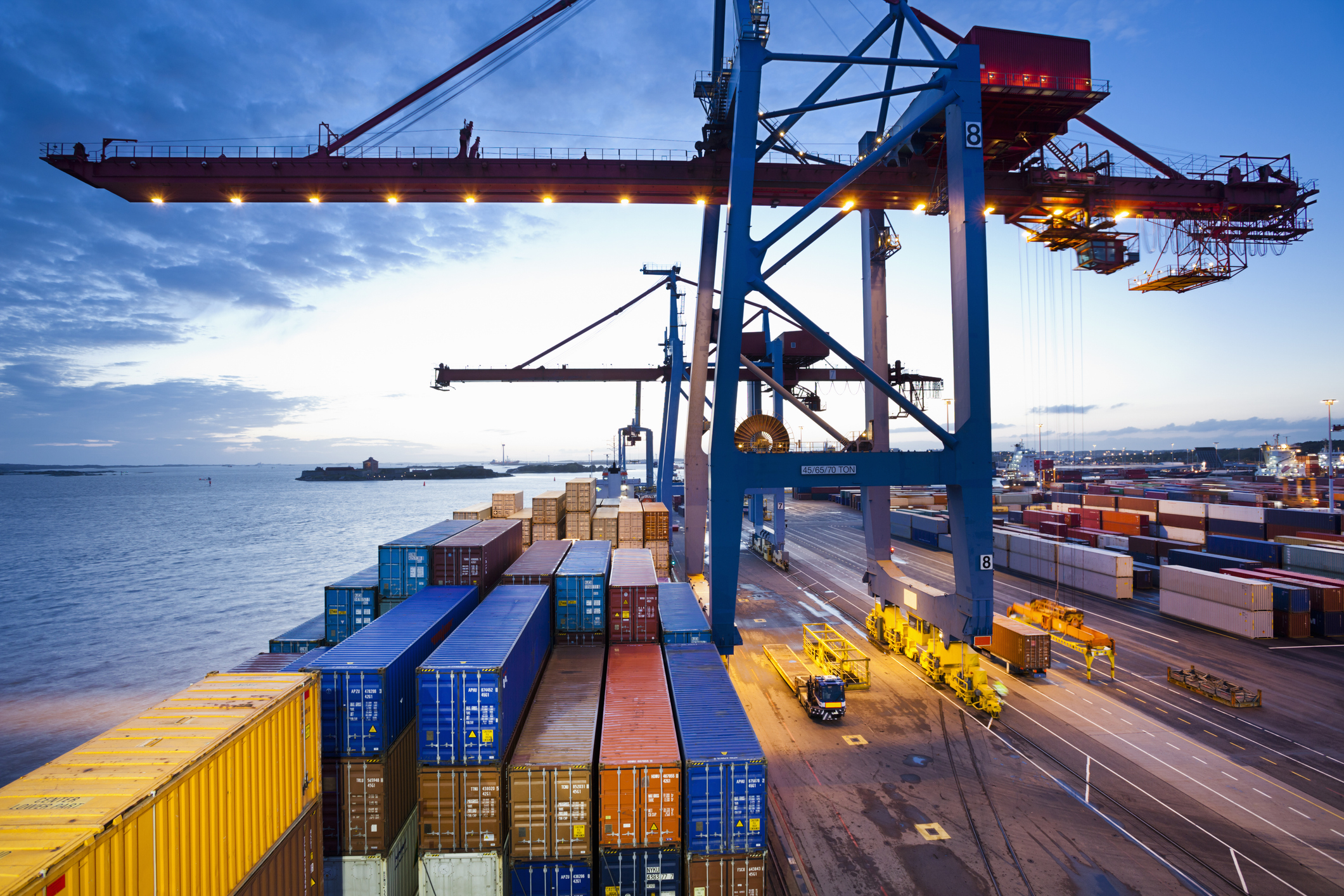 Kranar flyttar containrar vid en hamnterminal nära vattnet, med lastbilar och staplad last synliga på kajen.