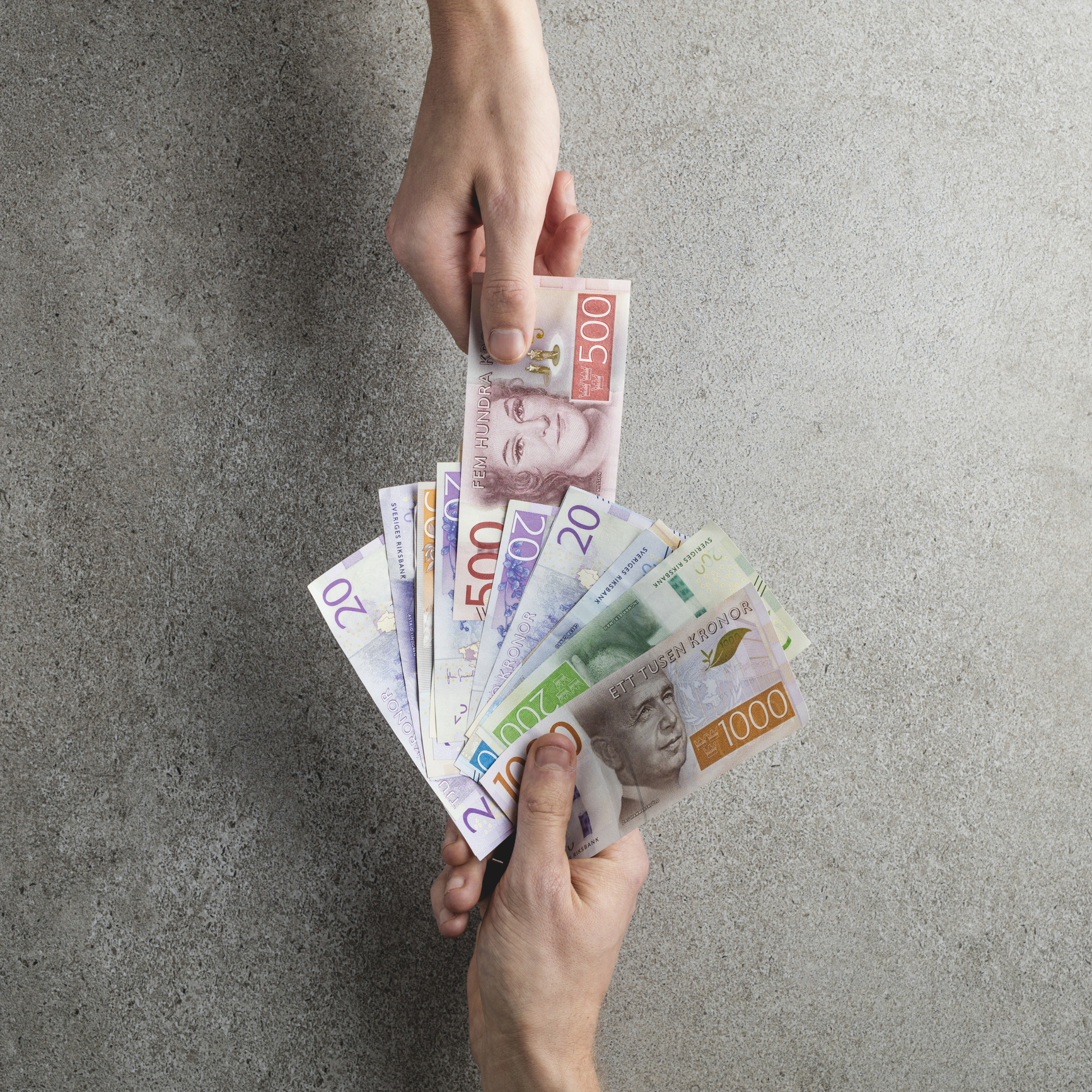 Två händer byter svenska sedlar i valörerna 20, 50, 100, 200, 500 och 1000 kronor på en grå yta.
