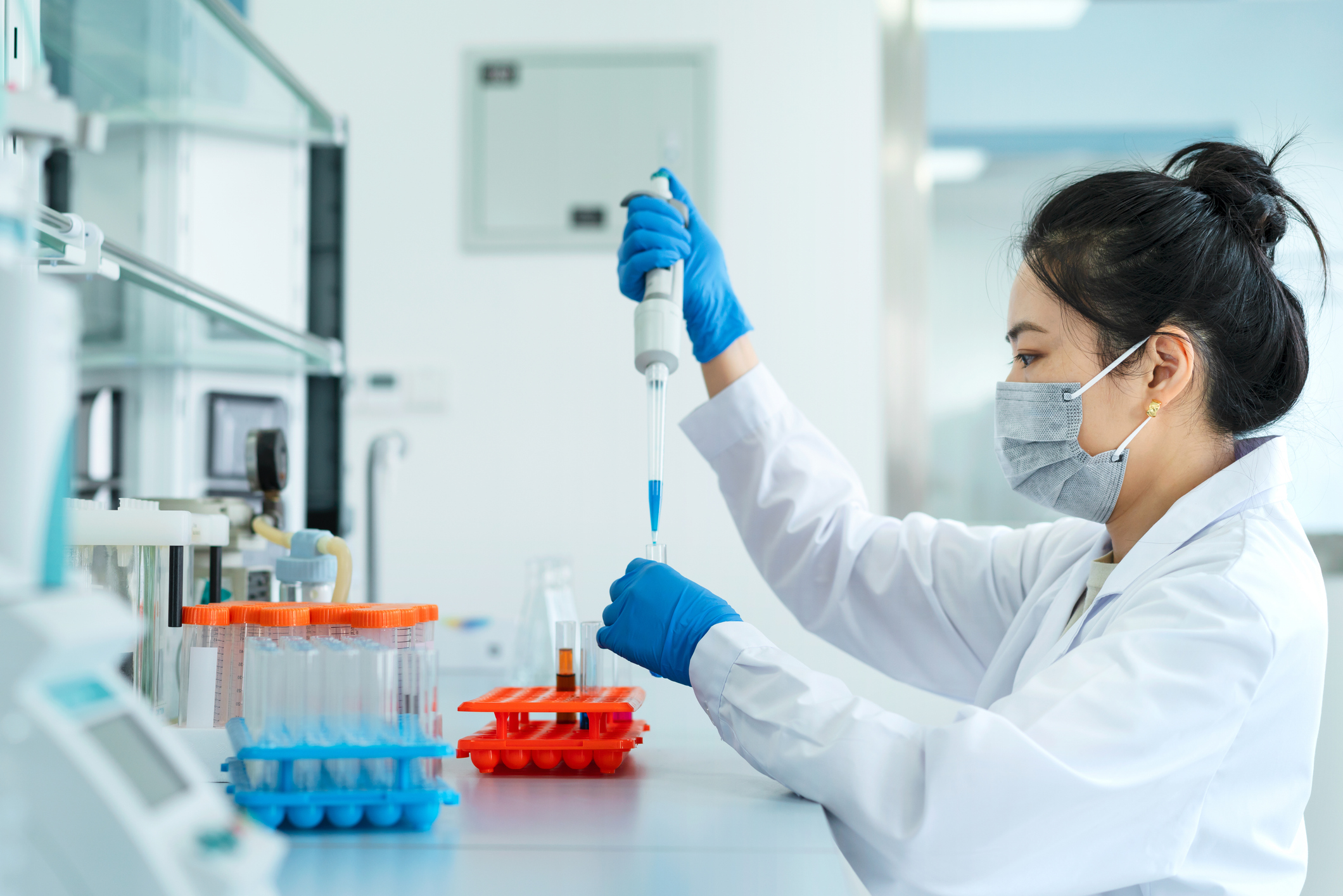 En forskare med labbrock och mask använder en pipett för att föra över vätska till provrör, omgiven av laboratorieutrustning.