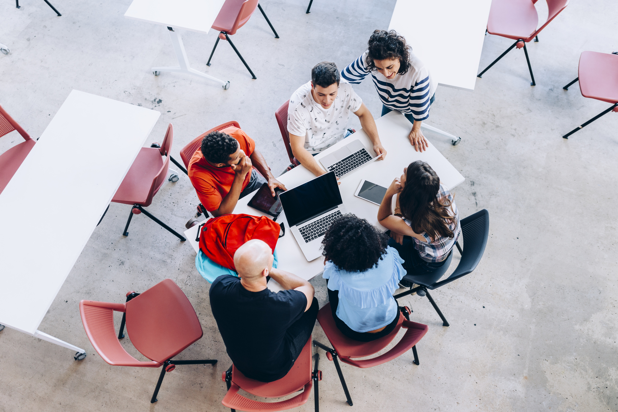 Fem personer sitter runt ett bord med datorer och surfplattor och arbetar tillsammans i ett rum med stolar och bord.