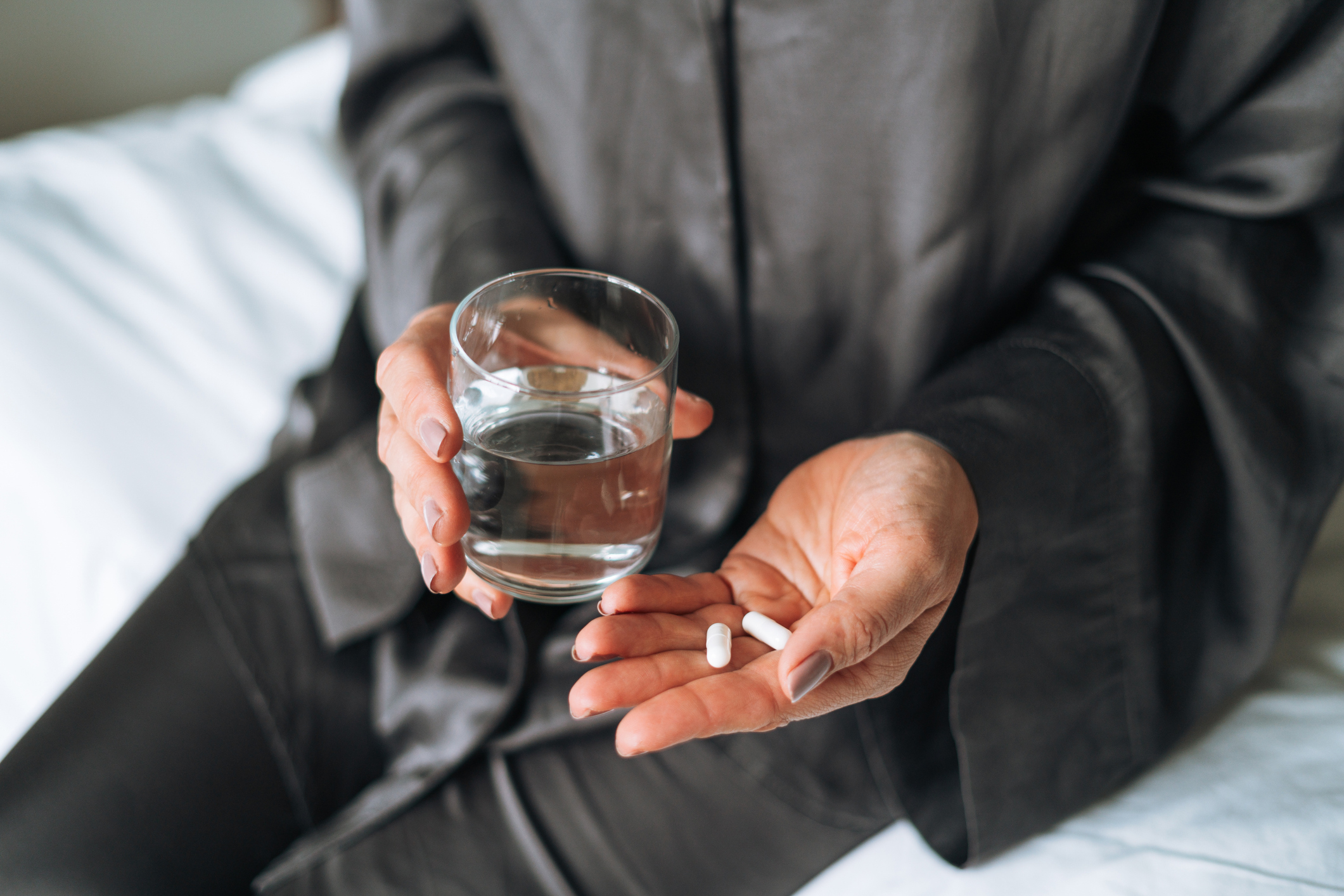 En person sitter på en säng med ett glas vatten i ena handen och ett vitt piller i den andra, klädd i mörka kläder.