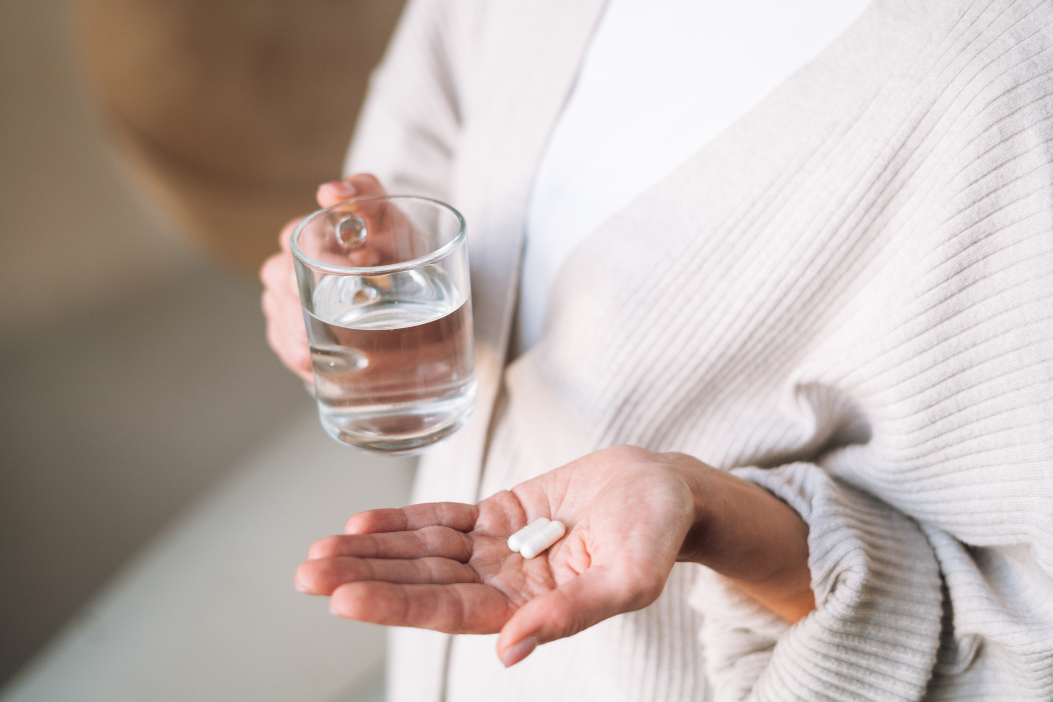 Person håller ett glas vatten i ena handen och två vita tabletter i den andra, iförd en ljus kofta.