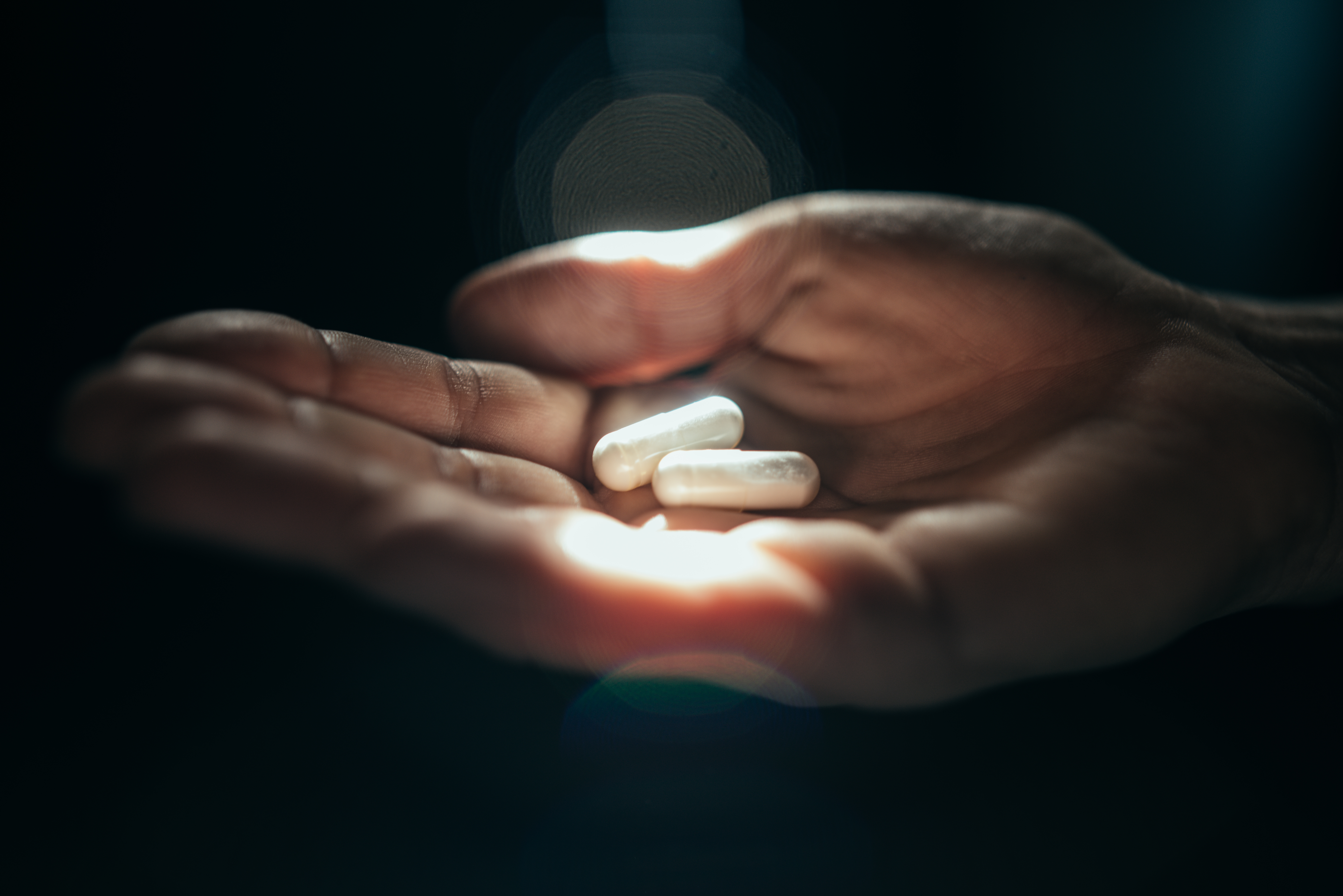 En öppen hand håller två vita kapslar, mot en mörk bakgrund med ljus riktat på kapslarna.