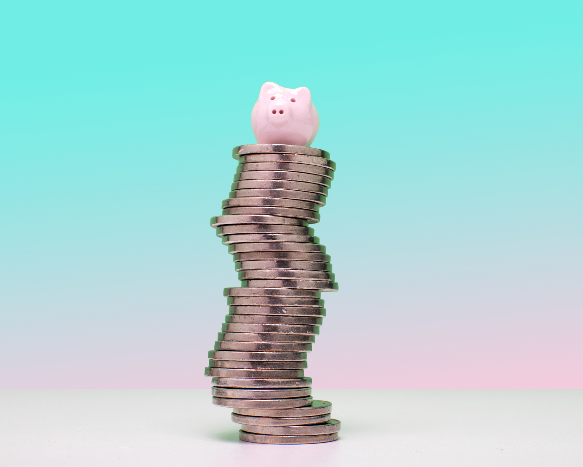 En rosa spargris står på en hög stapel mynt framför en bakgrund i turkos och rosa toning.