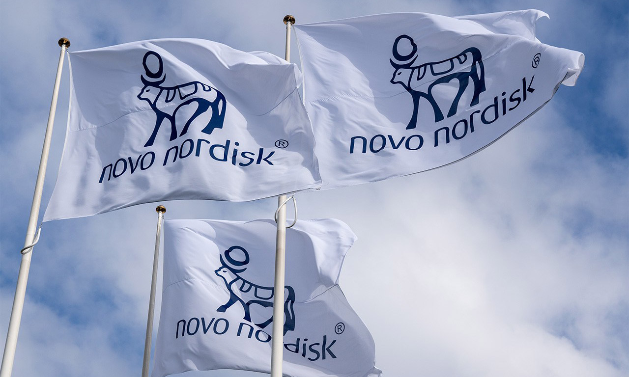 Tre vita flaggor med blå Novo Nordisk-logotyp och namn hissade på flaggstänger mot en molnig himmel.