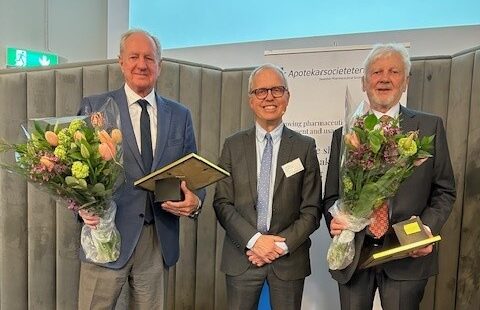 Tre män i kostym står inomhus; de till vänster och höger håller inramade diplom och blombuketter. En skärm och en banner syns i bakgrunden.