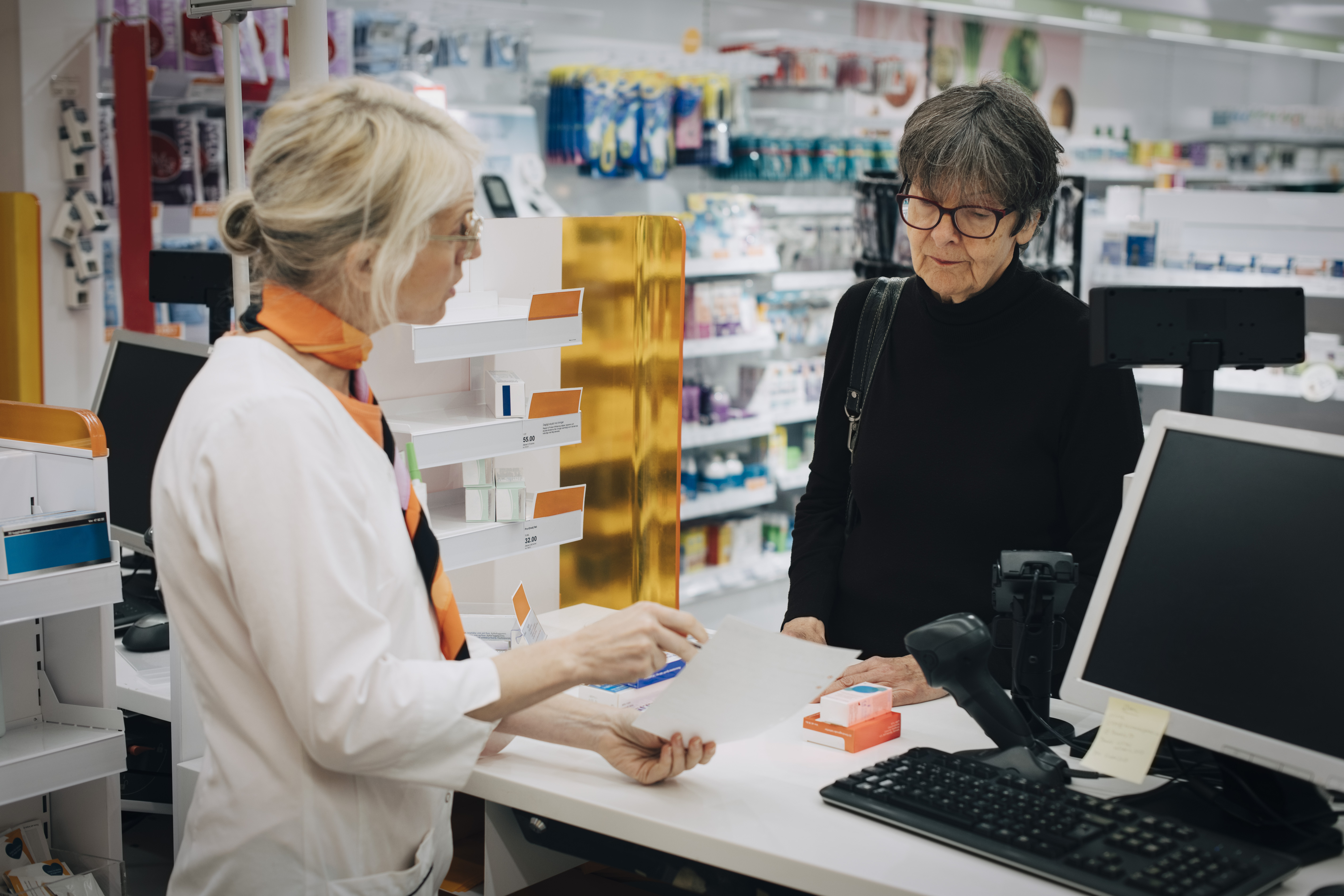 En farmaceut samtalar med en äldre kvinna vid ett apoteksställe om papper och medicin. Hyllor med produkter syns i bakgrunden.