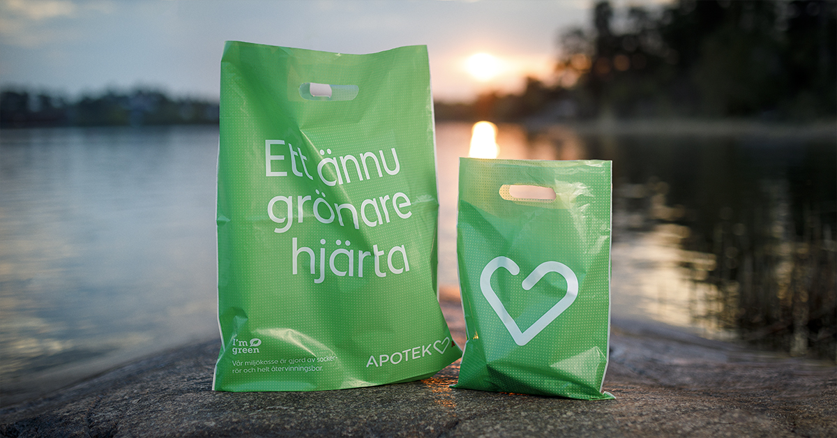 Två gröna plastpåsar med vit text och hjärtsymbol ligger på en sten vid vatten i solnedgången.