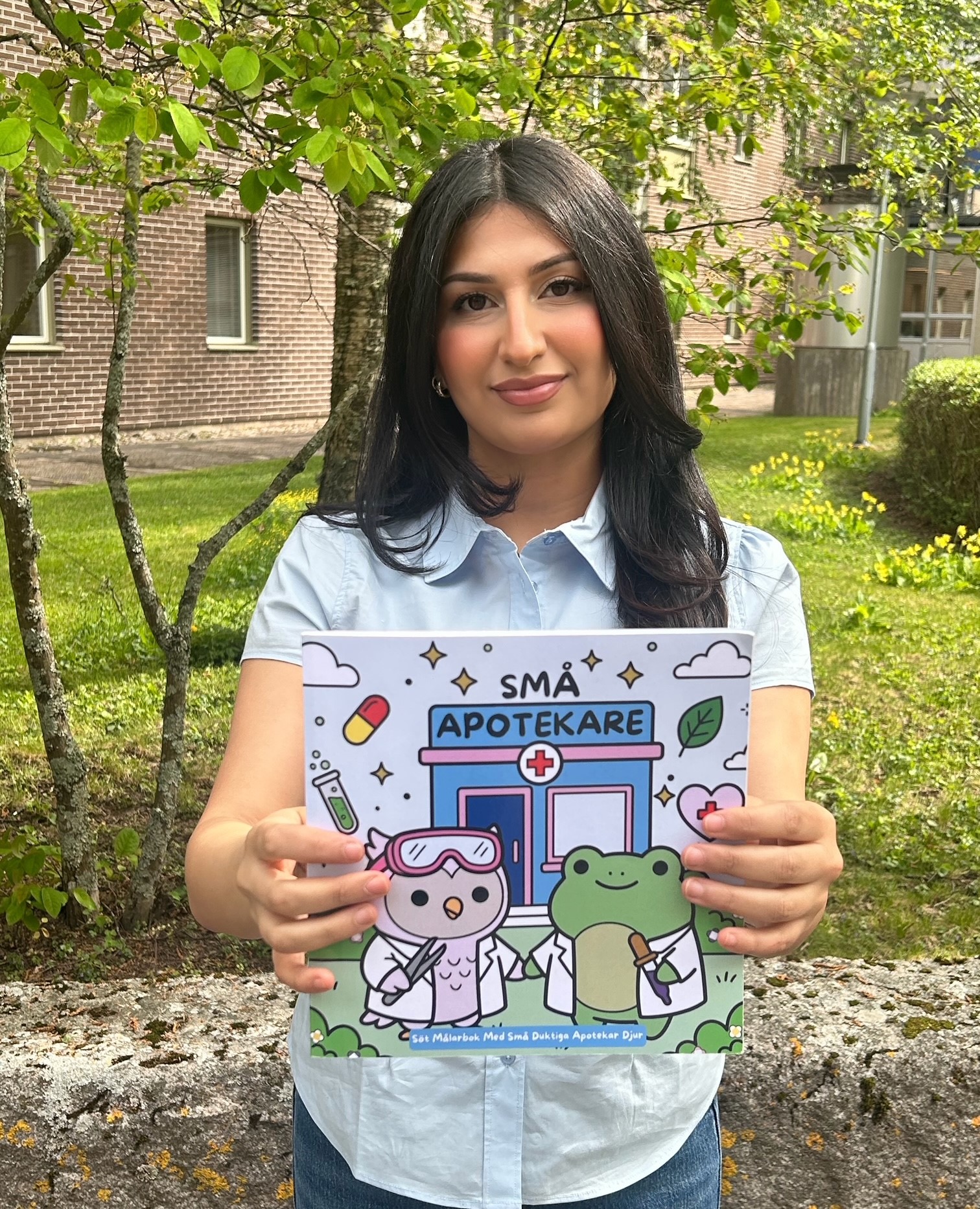 En kvinna står utomhus och håller upp en barnbok med titeln "Små Apotekare" och tecknade djur på omslaget.