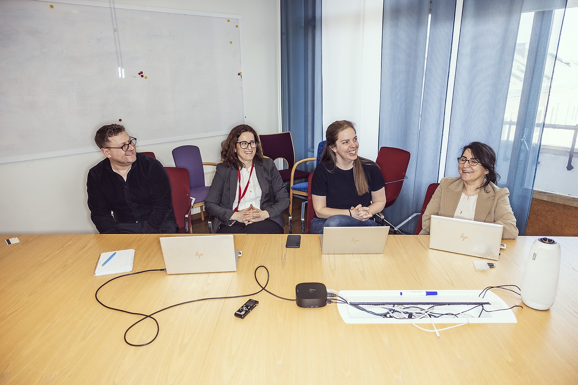 Fyra personer sitter vid ett konferensbord med bärbara datorer och anteckningsblock i ett mötesrum med whiteboard och blå gardiner.