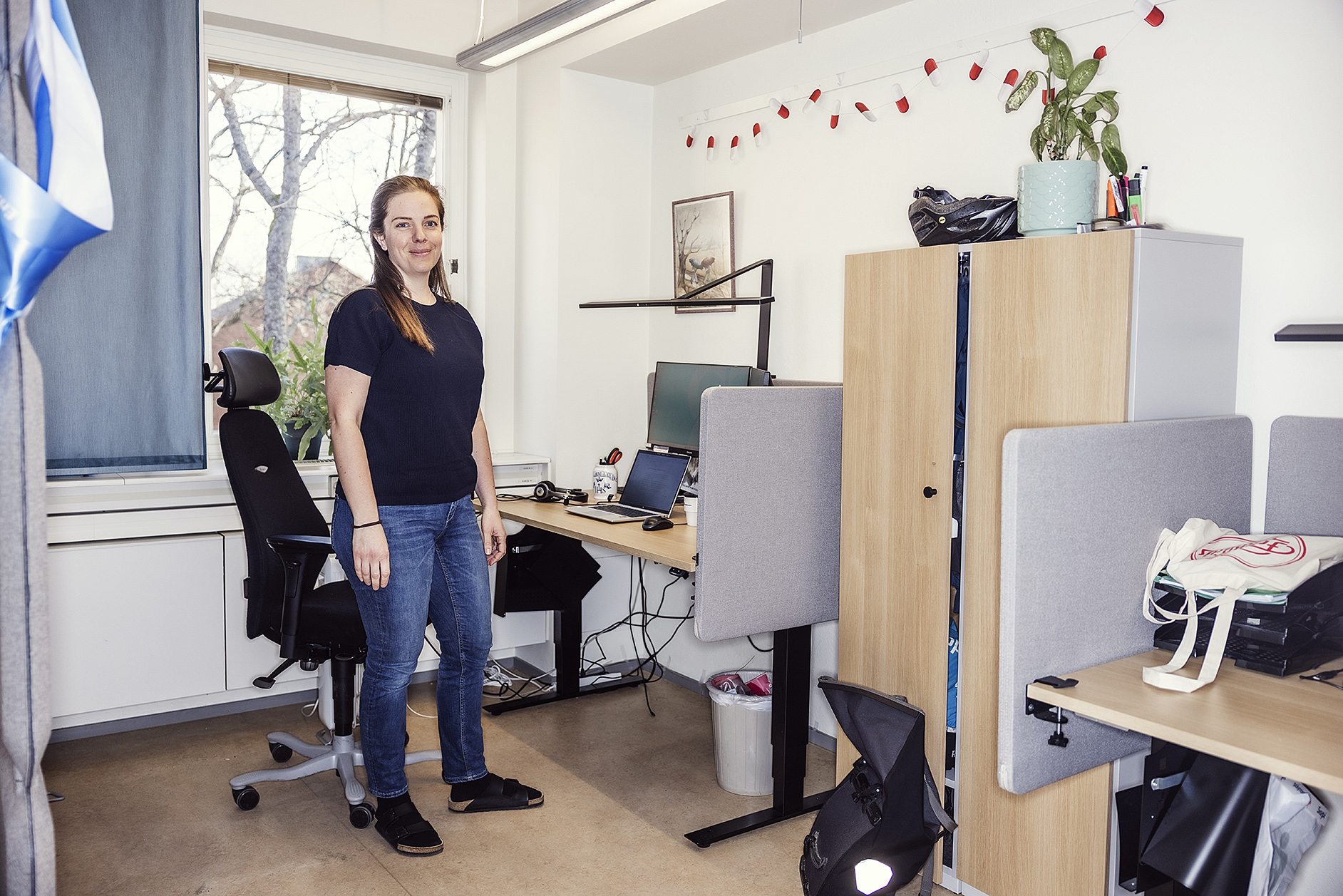 En kvinna står i ett kontor bredvid sitt skrivbord med en dator, kontorsstol och personliga saker. Två skrivbord, skåp och ett fönster finns i rummet.