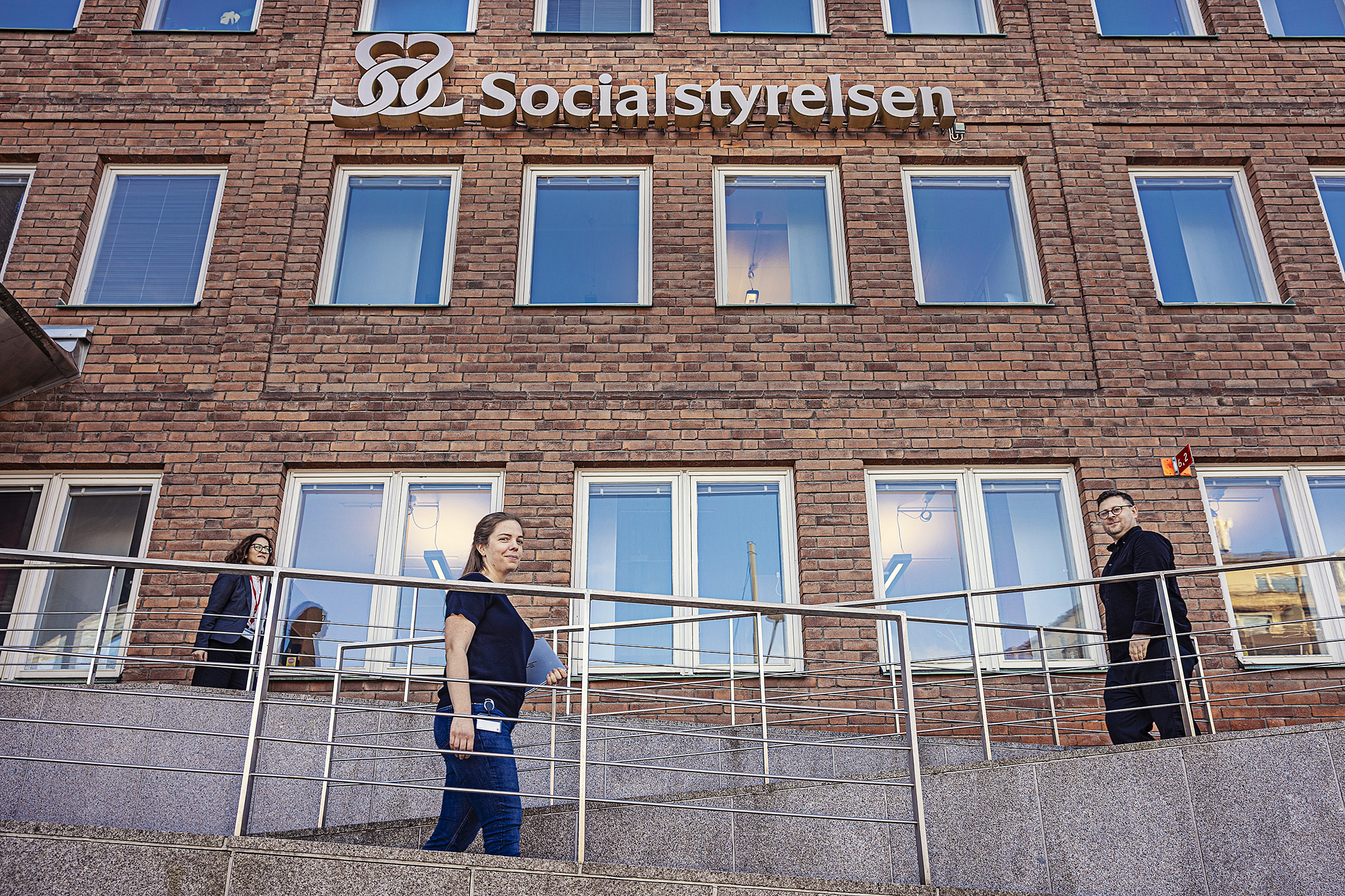 Tre personer går utanför en tegelbyggnad med skylten "Socialstyrelsen" ovanför ingången.