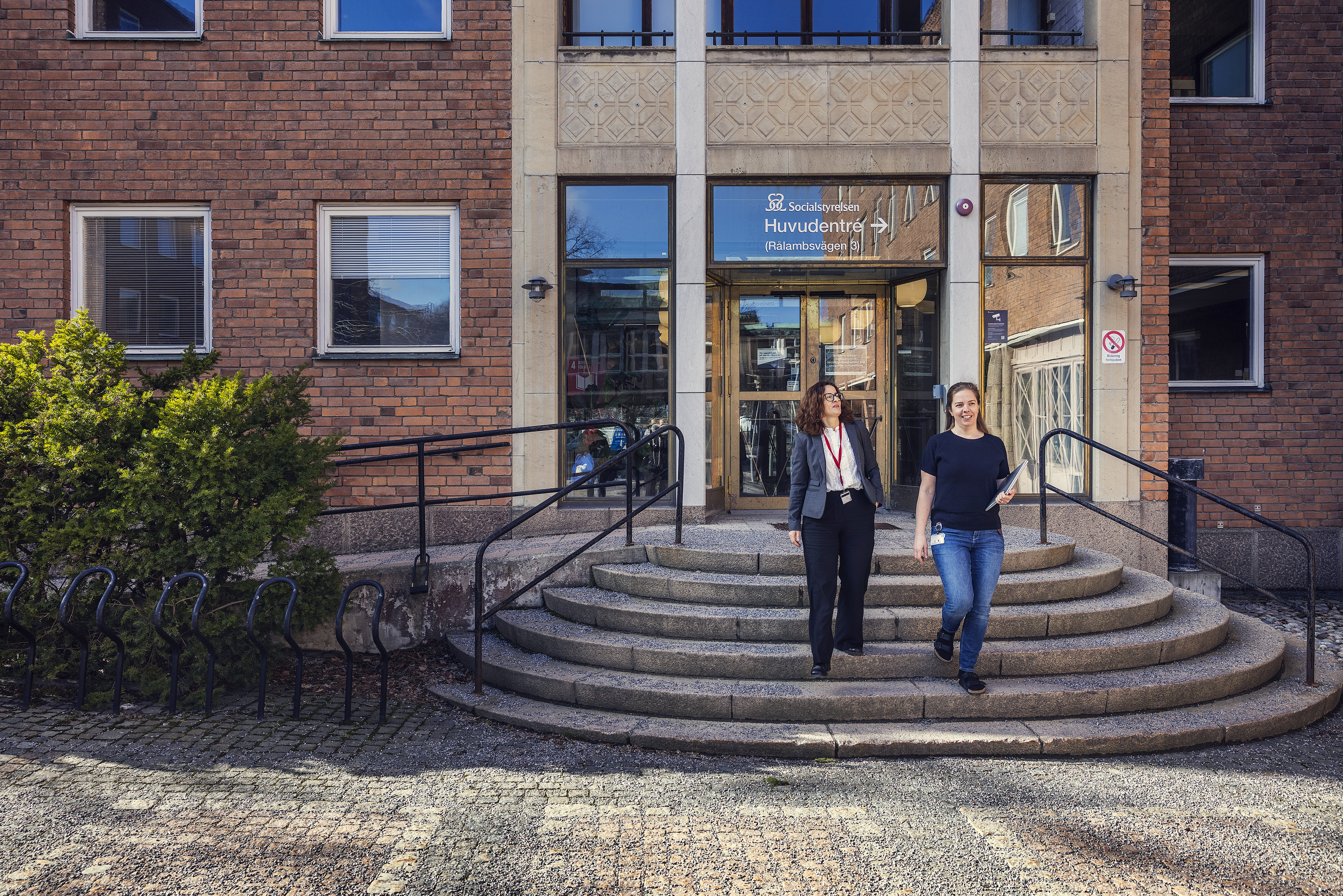 Två personer går nerför trappan framför en tegelbyggnad med glasdörrar och skylt ovanför ingången.