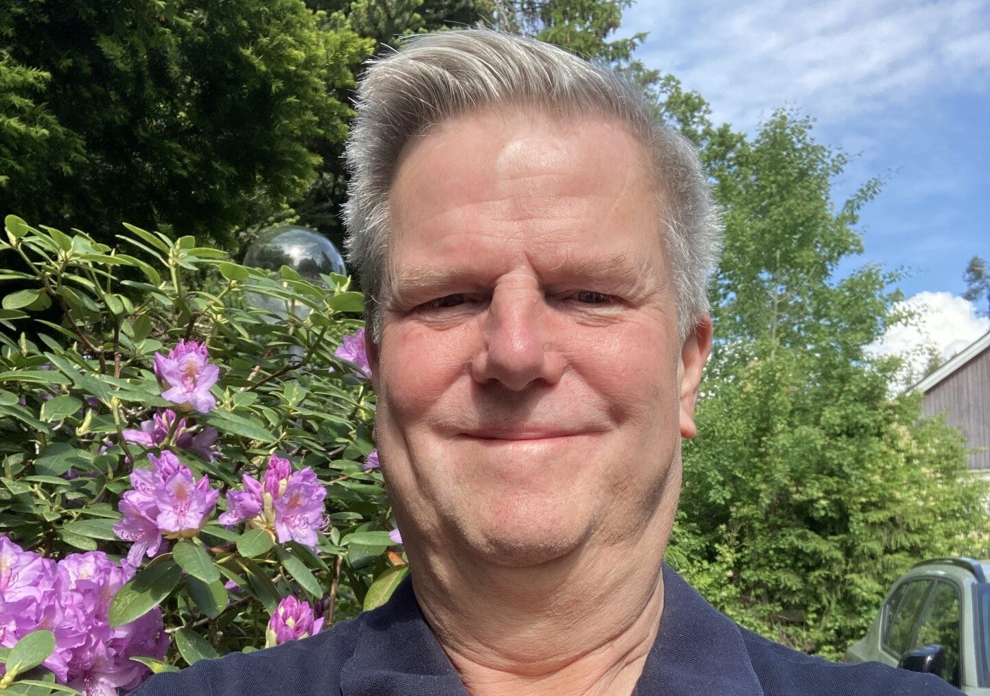 En man tar en selfie utomhus med lila blommor och gröna träd i bakgrunden under en solig dag.