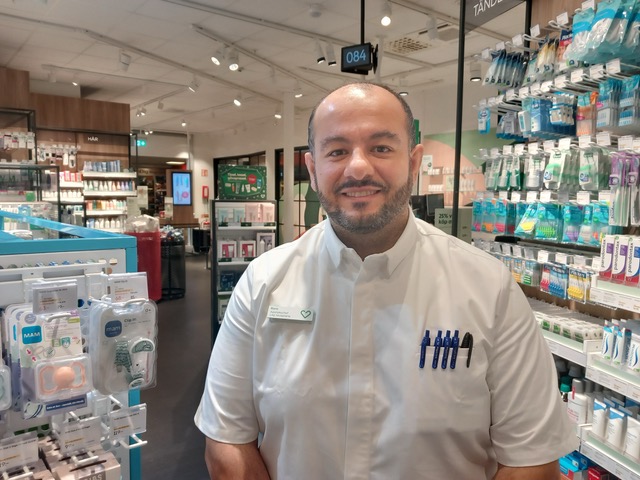 En farmaceut i vit rock står i ett apotek, omgiven av hyllor med produkter för hälsa och personlig vård.