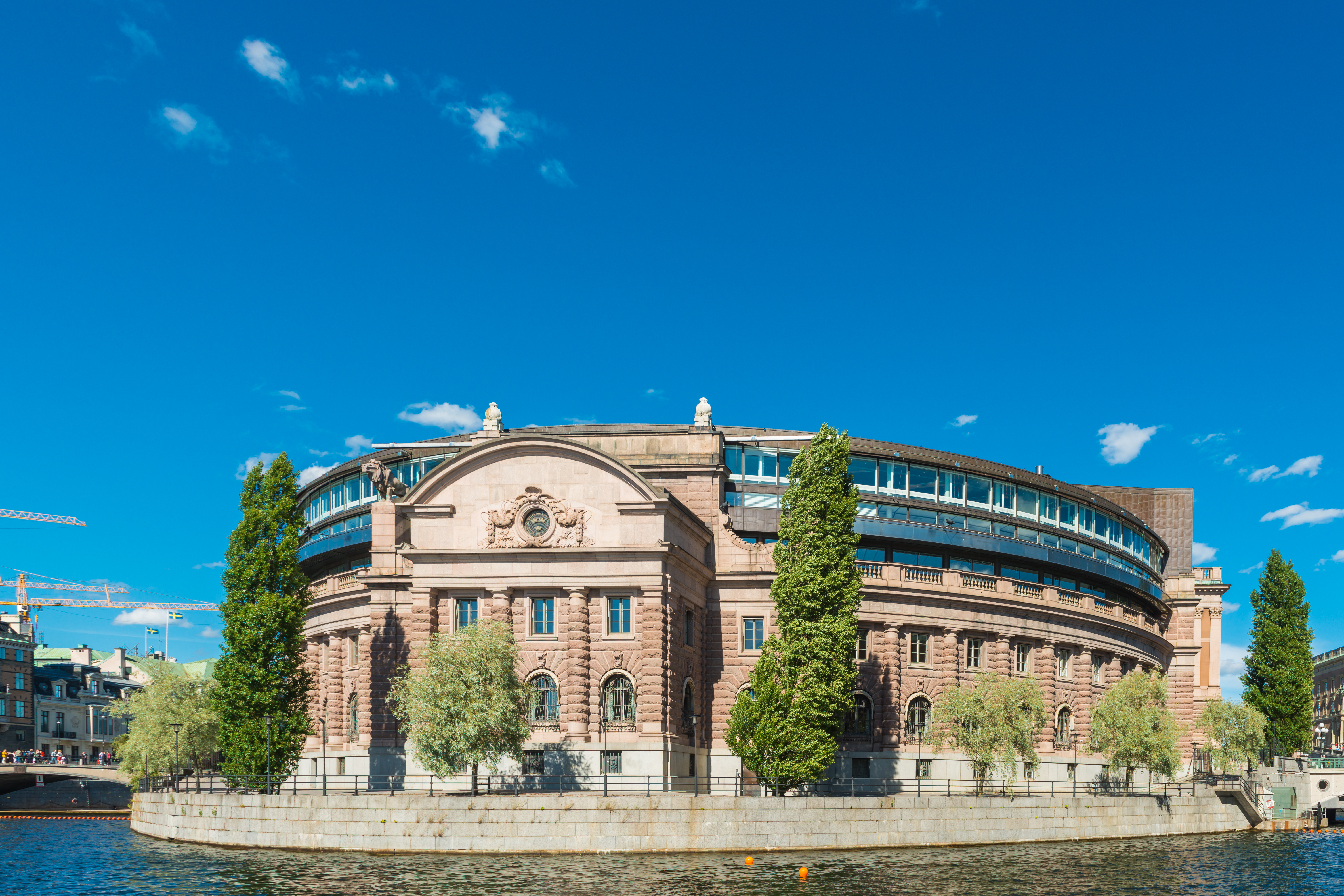 En stor historisk byggnad med rundad fasad och en modern glasdel, belägen vid vattnet under en blå himmel med träd i förgrunden.
