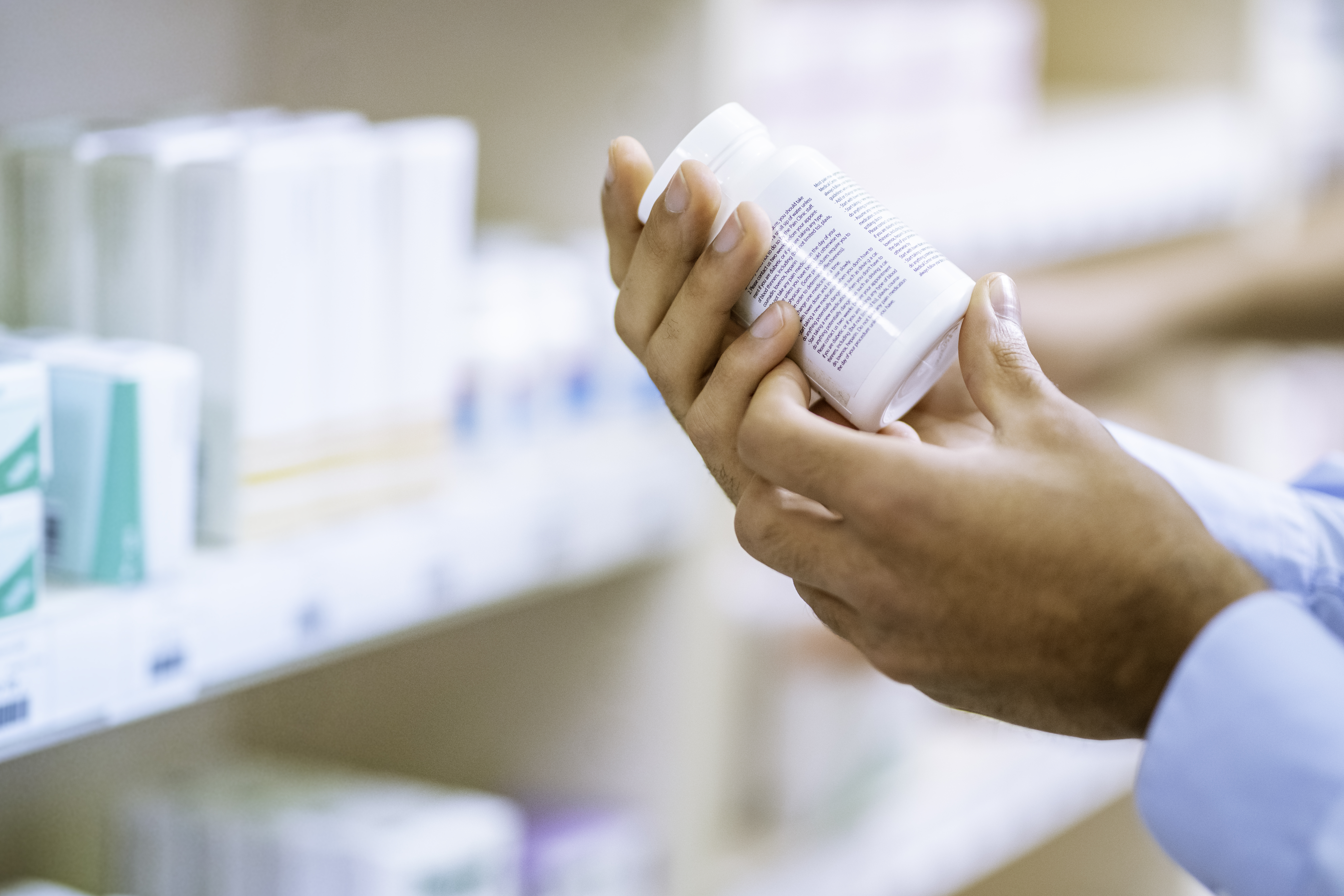 En person håller i en vit tablettflaska och läser på etiketten i ett apotek, med hyllor fyllda med medicinförpackningar i bakgrunden.