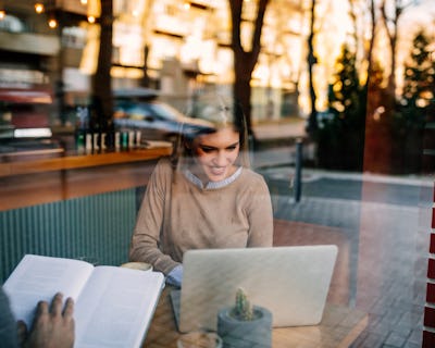 Kvinna som läser en bok och använder en bärbar dator på ett café.