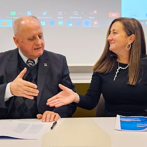 Två personer engagerade sig i en diskussion vid ett möte med ett videokonferensgränssnitt som visas på en skärm i bakgrunden.