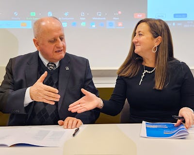 Två personer engagerade sig i en diskussion vid ett möte med ett videokonferensgränssnitt som visas på en skärm i bakgrunden.