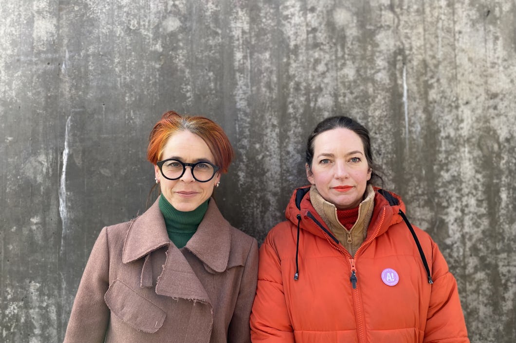Två kvinnor står framför en betongvägg, den ena med glasögon och brun rock, den andra i röd jacka med nål.