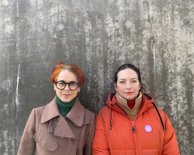 Två kvinnor står framför en betongvägg, den ena med glasögon och brun rock, den andra i röd jacka med nål.
